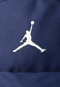 Tengeri textúrájú anyag, amelyen egy fehér sziluett látható, amely egy ugró alakot ábrázol, közismert nevén a Jumpman logó, hangsúlyosan megjelenítve az anyagon.