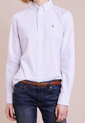 Osoba ubrana w białą koszulę na guziki w pionowe, jasnoniebieskie paski, wpuszczoną w ciemnoniebieskie dżinsy z brązowym paskiem.