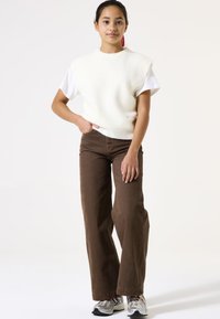 Pull en maille côtelée crème sans manches sur un t-shirt blanc, associé à un pantalon large marron et des baskets grises.