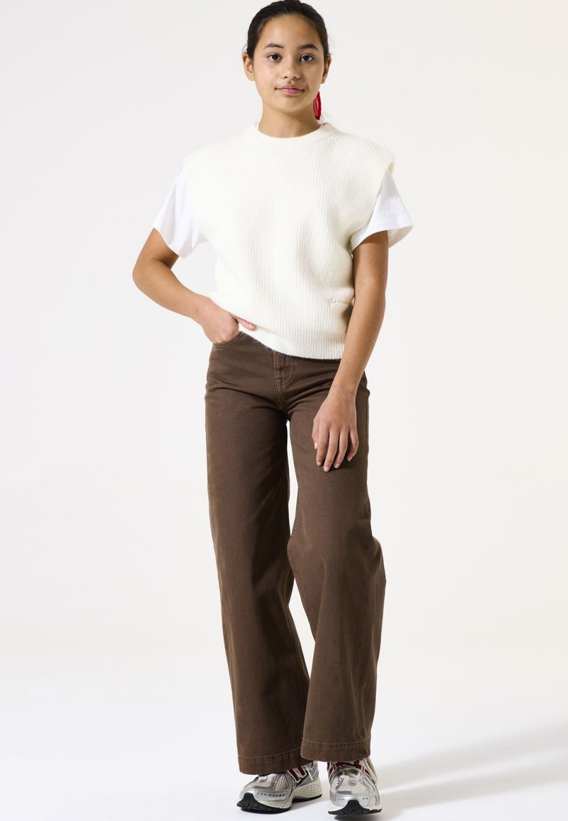 Pull en maille côtelée crème sans manches sur un t-shirt blanc, associé à un pantalon large marron et des baskets grises.