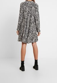 Abito leopardato nero e beige con maniche lunghe, design ampio e a strati, e colletto con bottoni. Indossato con stivali neri con tacco alla caviglia.