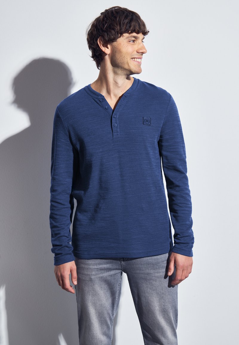 Man die glimlacht en naar rechts kijkt, gekleed in een blauw lange mouwen Henley shirt en lichtgrijze jeans, staande tegen een effen witte muur.