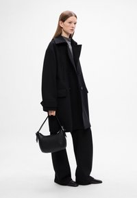 Cappotto oversize nero con ampio colletto, doppi bottoni e tasche anteriori, abbinato a pantaloni larghi neri e una piccola borsa nera a tracolla.