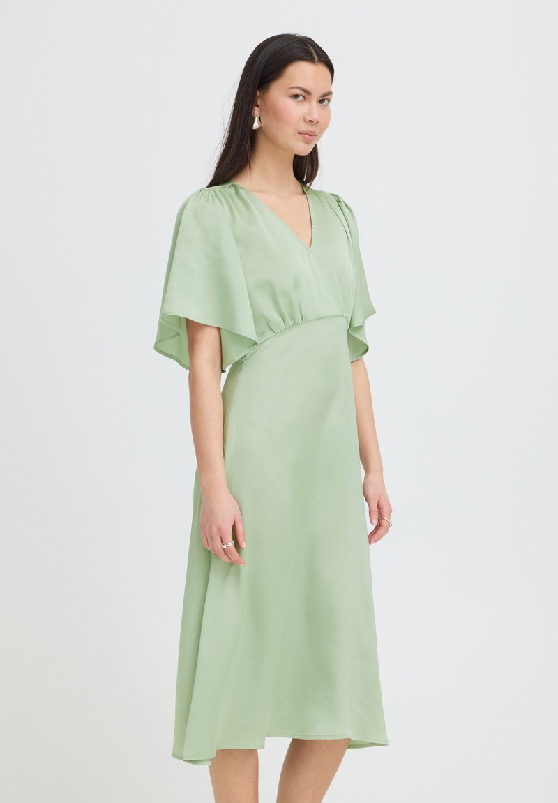 Femme portant une robe midi vert clair avec des manches volantes et un col en V, debout devant un fond gris clair uni.