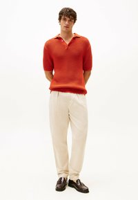 Polo shirt a maglia rossa con texture, maniche corte, abbinato a pantaloni beige chiaro e mocassini marrone scuro, in piedi contro uno sfondo semplice.
