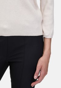 Helles beiges, geripptes Sweatshirt mit abgerundetem Saum und 3/4-Ärmeln, kombiniert mit schwarzen, figurbetonten Hosen. Hand ruht sanft auf dem Kleidungsstück.