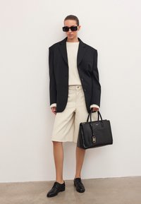 Donna che indossa un blazer nero oversize, un maglione beige e pantaloni corte, scarpe oxford nere, occhiali da sole, e porta una borsa nera con texture.