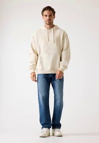 Beżowa bluza z kapturem z przednią kieszenią, zestawiona z niebieskimi jeansami i białymi trampkami. Materiał wydaje się miękki i teksturowany.