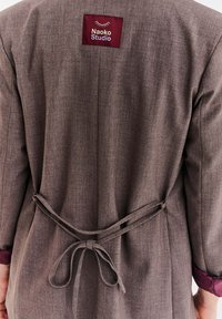 Brązowy szlafrok z tkaniny o fakturowanym wykończeniu, z bordową metką z napisem "Naoko Studio" oraz paskiem przewiązywanym w talii.