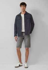 Marineblauwe katoenen jas met twee borstzakken, gedragen over een wit T-shirt, gecombineerd met grijze denim shorts en witte sneakers met zwarte accenten.