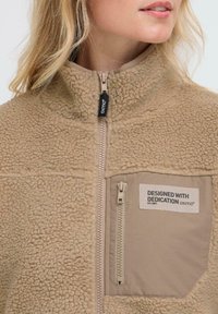 Veste en polaire beige avec col haut, fermeture éclair à l'avant, poche poitrine zippée et patch portant la mention "Conçu avec dévouement oxmo®".