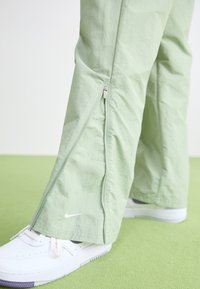 Pantalones ligeros de color verde claro con cremalleras laterales y un sutil logo de Nike. Combinados con zapatillas blancas, en un fondo verde.