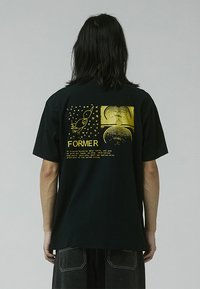 Camiseta de algodón negra con estampados gráficos dorados en la parte posterior, incluyendo un cohete, estrellas y la palabra "FORMER" con texto adicional.
