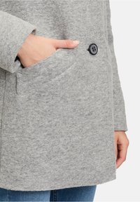 Cappotto di lana grigio con finitura testurizzata, dotato di chiusura a bottoni, tasca laterale e taglio dritto. Mano visibile nella tasca.