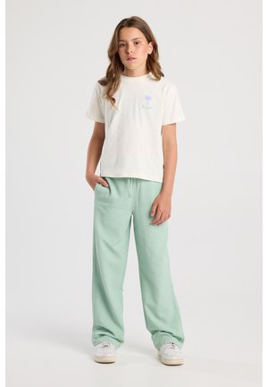 T-shirt crème clair avec un graphique de palmier et un pantalon en mint vert texturé. Les deux pièces sont décontractées, avec une coupe ample et un design simple. Des baskets blanches complètent le look.