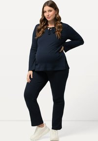 Navyblå graviditetsblus med lång ärm och knytkrage, ihop med matchande åtsittande byxor. Både i mjukt, текстurerat tyg. Vita sneakers.