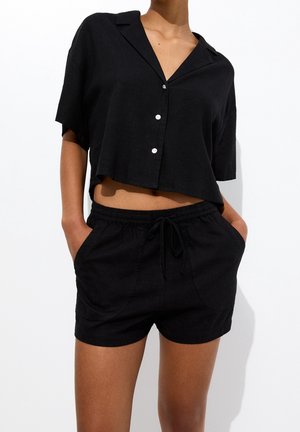 Mujer con camisa negra de botones corta con cuello y pantalones cortos negros con cordón, de pie con las manos en los bolsillos sobre fondo blanco.