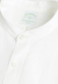 Polo bianco slim-fit con colletto a fascia, dotato di una chiusura con un singolo bottone e un’etichetta verde con la scritta "Brooks Brothers 1818."