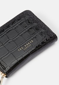 Cartera negra brillante con textura de cocodrilo, presenta la marca dorada "TED BAKER LONDON" y un cierre con cremallera y tirador de metal.