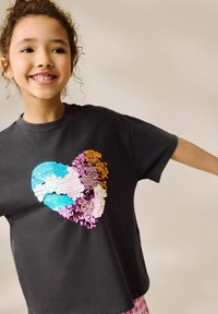 Schwarzes T-Shirt mit kurzen Ärmeln, das ein Herzdesign aus mehrfarbigen Pailletten in Blau-, Pink- und Goldtönen zeigt.