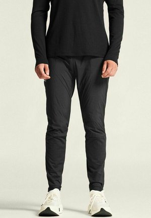 Moitié inférieure d'une personne portant un pantalon noir, une chemise à manches longues noire et des baskets blanches, se tenant devant un fond uni.