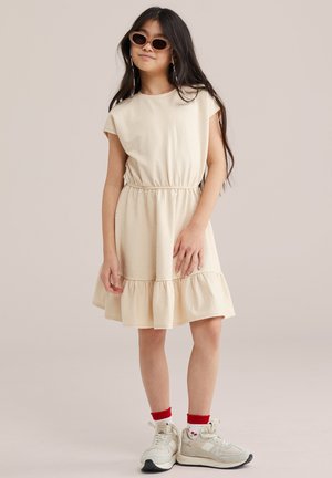 Hosszú sötét hajú lány bézs csíkos ruhában, piros díszítésű zokniban, fehér tornacipőben és barna napszemüvegben, egyszínű háttér előtt állva.