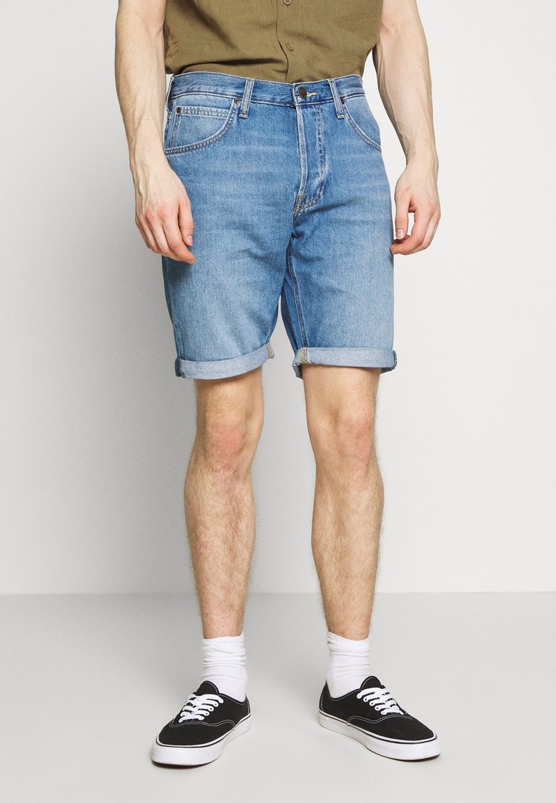 Homme portant un short en denim bleu clair retroussé, des chaussettes blanches, des baskets noires et une chemise vert olive sur un fond clair uni.