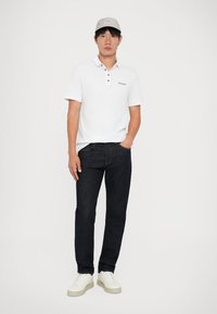 Hvid polo-shirt med knapkrave, parret med mørke denimjeans og hvide sneakers. Personen bærer en lysegrå kasket. Ensfarvet baggrund.