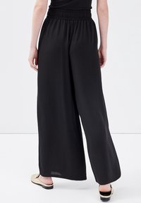 Pantalon noir à jambes larges avec une ceinture élastique, confectionné dans un matériau léger, présentant une texture lisse sans motifs visibles.
