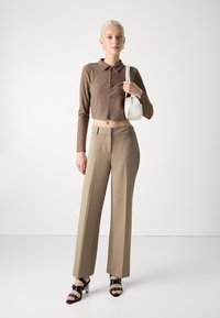 Hnědý žebrovaný polo crop top s dlouhým rukávem v kombinaci s béžovými vysokými širokými kalhotami a černými sandály na podpatku s pásky, držící bílou tašku.
