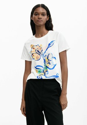 Naine, kes kannab valget lühikeste varrukatega T-särki suure sinise ja beeži lillemustriga, mustade pükstega, seisab valge tausta ees.