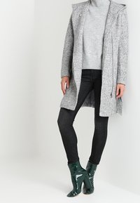 Grå texturerad kappa med huva, kombinerad med en ljusgrå polotröja och svarta jeans. Grönt blanka ankelskor med klack.