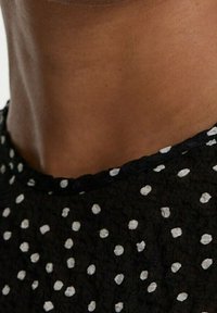 Schwarzer Stoff mit weißem Punktmuster, strukturierte Oberfläche, runder Auschnitt mit kontrastierendem schwarzen Rand, zeigt einen Teil des Nackens und der Schulter.