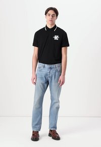Schwarzes Polohemd mit weißen Kragenakzenten und Logo, kombiniert mit hellblauen Jeans und braunen Schuhen. Modell steht vor einem einfarbigen Hintergrund.