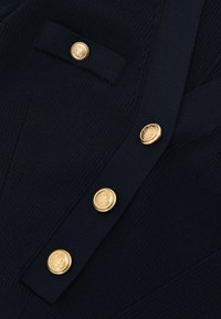 Marineblauw geribd gebreid kledingstuk met gouden ronde knopen met ingewikkeld detail. Inclusief een zakklep met een bijpassende knop.
