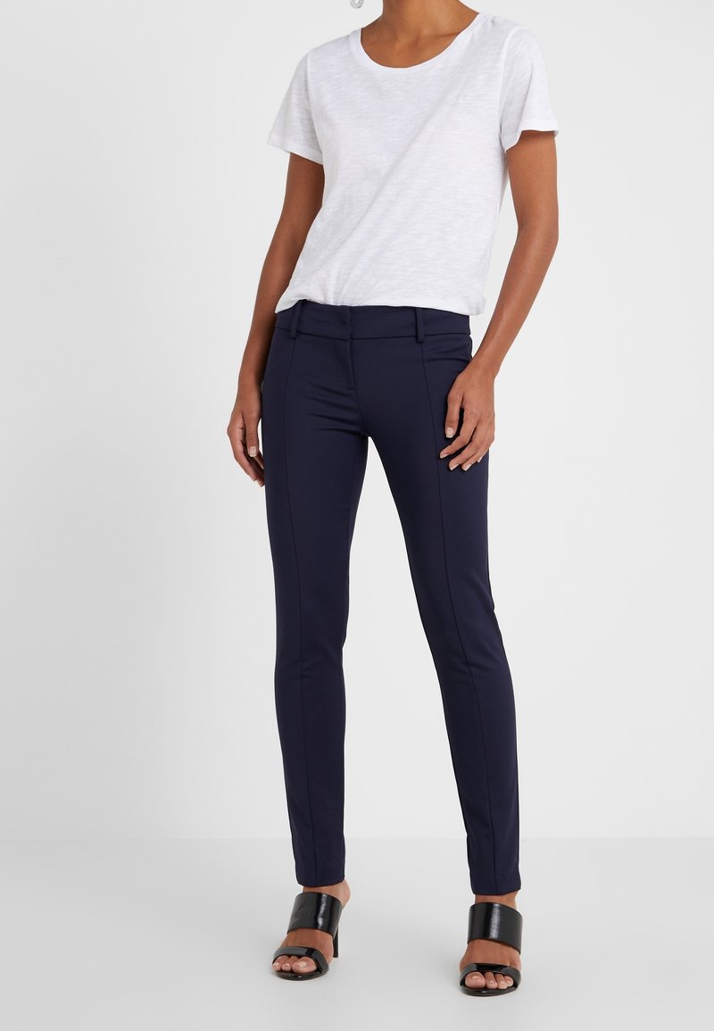 Femme portant un pantalon slim-fit bleu marine, un t-shirt blanc à manches courtes et des sandales à talons ouverts noires, se tenant devant un fond uni.