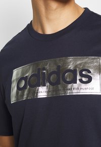 Camiseta de algodón azul marino con un gran logo metálico plateado de "adidas" con texto, cuello redondo y mangas cortas.