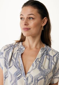 Blusa a maniche corte in tessuto crema con un motivo geometrico blu navy. Dettagli notevoli includono una pattina con bottoni e una forma morbida e drappeggiata.