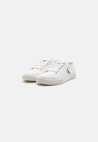 Witte canvas sneakers met een ronde neus, vetersluiting, een geribbelde rubberen zool en een klein zwart logo aan de zijkant.