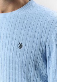 Lyseblå strikkegenser med kabelmønster, rund hals og en liten mørk logo brodert på brystet.