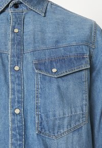 Ljusblå denimskjorta med spetsig krage, tryckknappar och bröstficka. Har en texturerad tygkvalitet och kontrasterande sömmar.