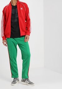 Röd zip-jacka med vita ränder, tillsammans med gröna byxor med röda sidosträck. Grå sneakers fulländar den sportiga stilen.