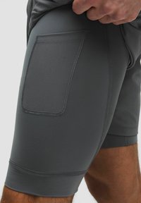 Gros plan sur un short de sport gris avec une poche latérale texturée et une finition en tissu lisse. Une main ajuste le tissu à la taille.