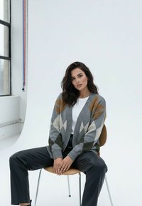 Kobieta o ciemnych włosach siedzi na drewnianym krześle, ubrana w szary kardigan we wzory, białą koszulę i czarne spodnie na jasnym, jednolitym tle.