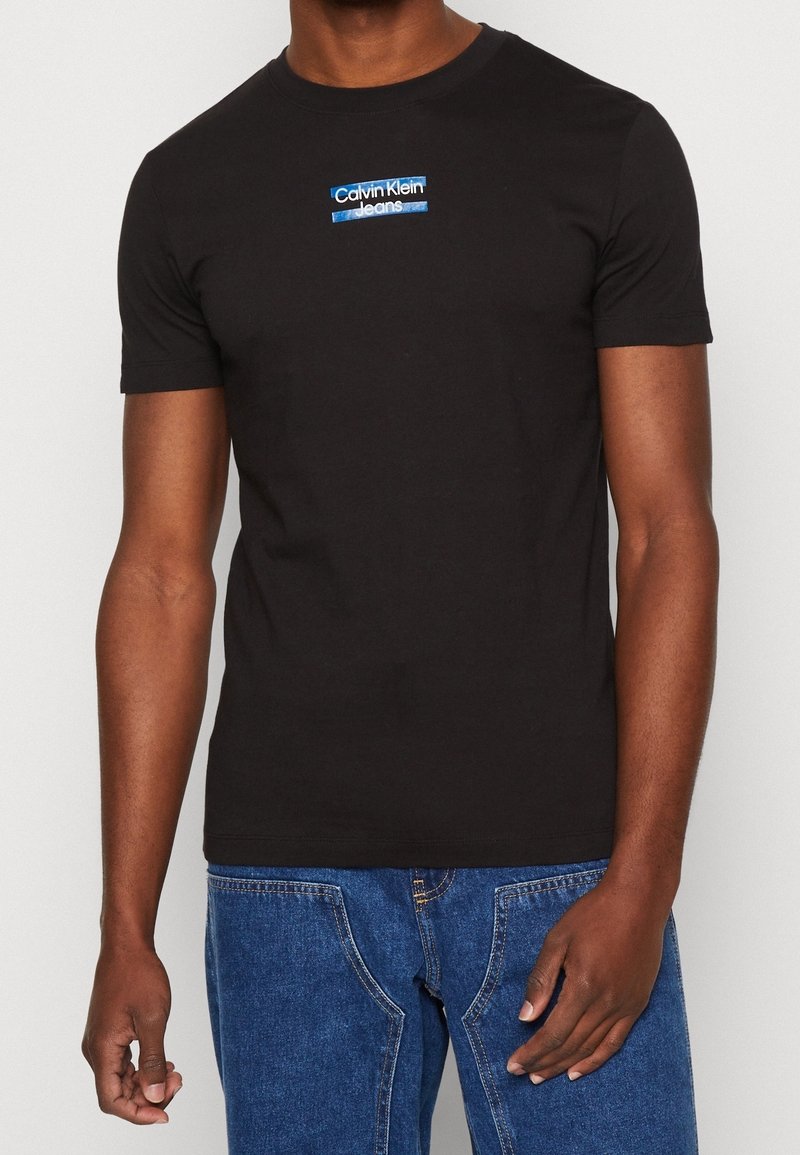Black cotton t-shirt with short sleeves, featuring a blue and white Calvin Klein Jeans logo on the chest. Classic crew neckline.