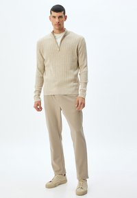 Jersey de canalé beige con cuello de cremallera, combinado con pantalones beige claros y zapatillas color tostado, creando un conjunto neutral cohesivo.
