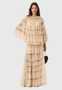 Krämfärgad klänning med flera lager i genomskinligt tyg, med volanger, hög krage och vida ärmar, tillsammans med en svart handväska.