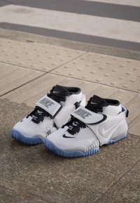 White athletic shoes with a textured surface, black accents, and a distinctive Nike strap across the front. Features a light blue rubber sole.