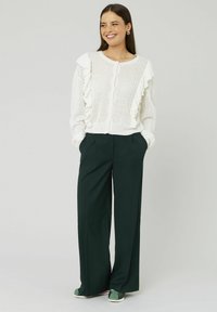 Cardigan blanc en tricot avec des bords ondulés, associé à un pantalon large vert foncé et des chaussures vertes à enfiler. Textures lisses et design superposé.
