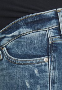 Jeans de denim azul con un lavado desgastado, que cuentan con un bolsillo frontal, costuras doradas y un detalle de botón plateado. La textura parece ligeramente usada.